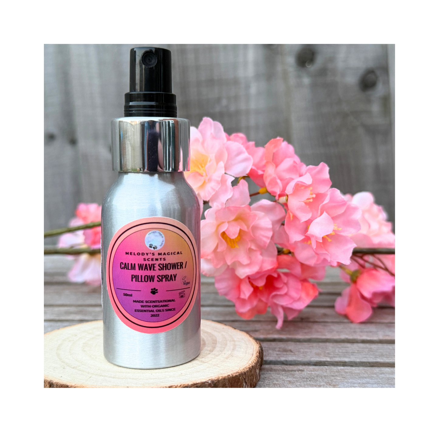 Bottle of 'Calm Wave Shower & Pillow Spray' with a pink label on a wooden surface with flowers in the background.
