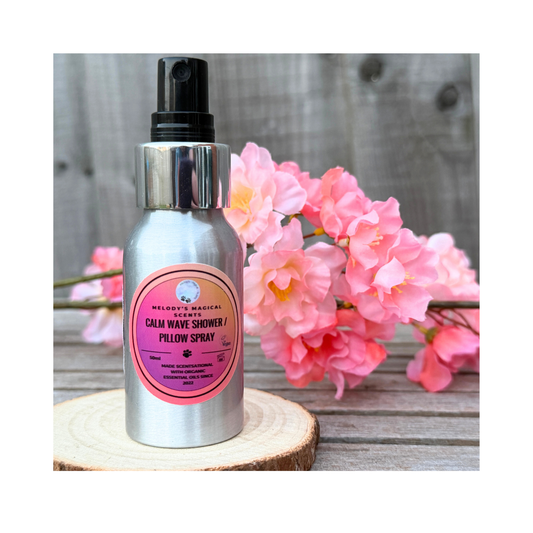 Bottle of 'Calm Wave Shower & Pillow Spray' with a pink label on a wooden surface with flowers in the background.