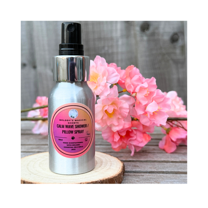 Bottle of 'Calm Wave Shower & Pillow Spray' with a pink label on a wooden surface with flowers in the background.