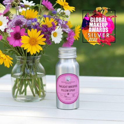 Bottle of 'Twilight Whispers Pillow Spray' with a pink label next to a vase of flowers on a white surface.