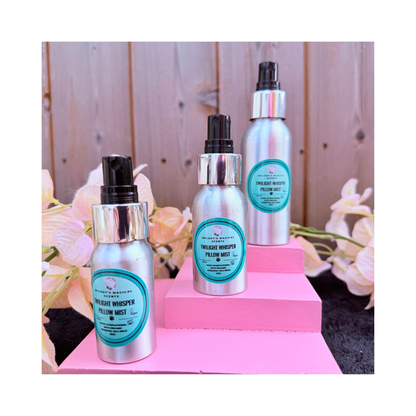 Three silver bottles with black nozzles on a pink stand against a wooden background. Twilight Whisper Pillow Mist trio - Jasmine and Chamomile aromatherapy pillow mist